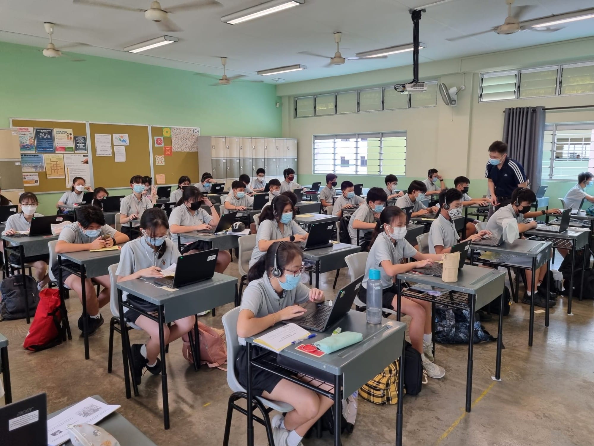 Students in masks sit at desks using laptops in a classroom while a teacher stands nearby.